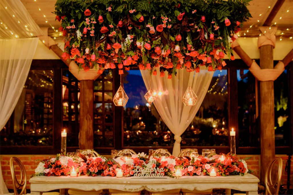 Mesa de novios decorada con flores rojas y velas, bajo un techo con follaje colgante y rosas, iluminación cálida y detalles románticos en un salón rústico elegante para boda, Sweetheart table decorated with red flowers and candles, under a hanging greenery and rose ceiling installation, with warm lighting in an elegant rustic wedding venue, Hacienda La Martina, La Martina Eventos