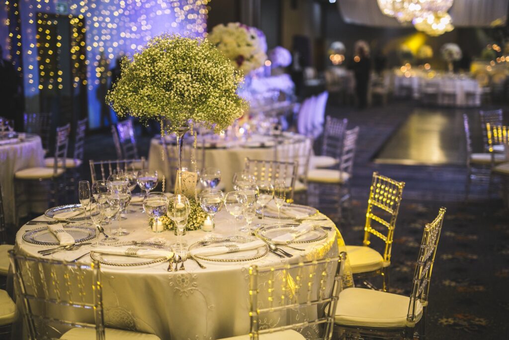 Centro de mesa de boda con arreglo alto de gypsophila en florero de cristal, mesa decorada con vajilla plateada y copas transparentes, ambientación elegante con luces cálidas y románticas
