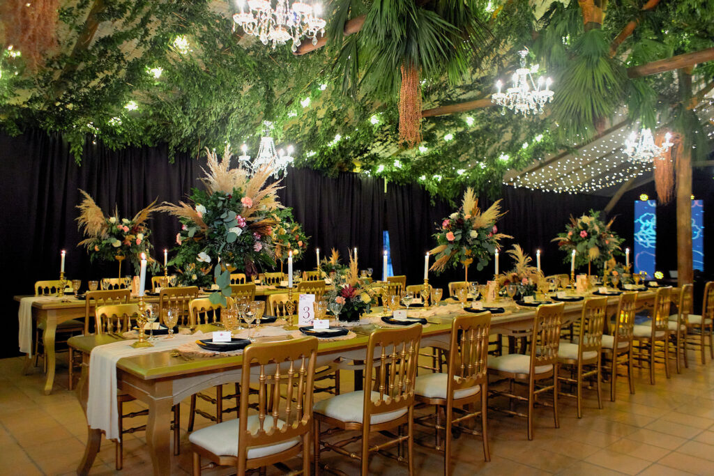 Centro de mesa de boda con flores, pampas y follaje verde en arreglos altos, mesas largas con sillas doradas, velas blancas y decoración de techo con follaje y luces cálidas