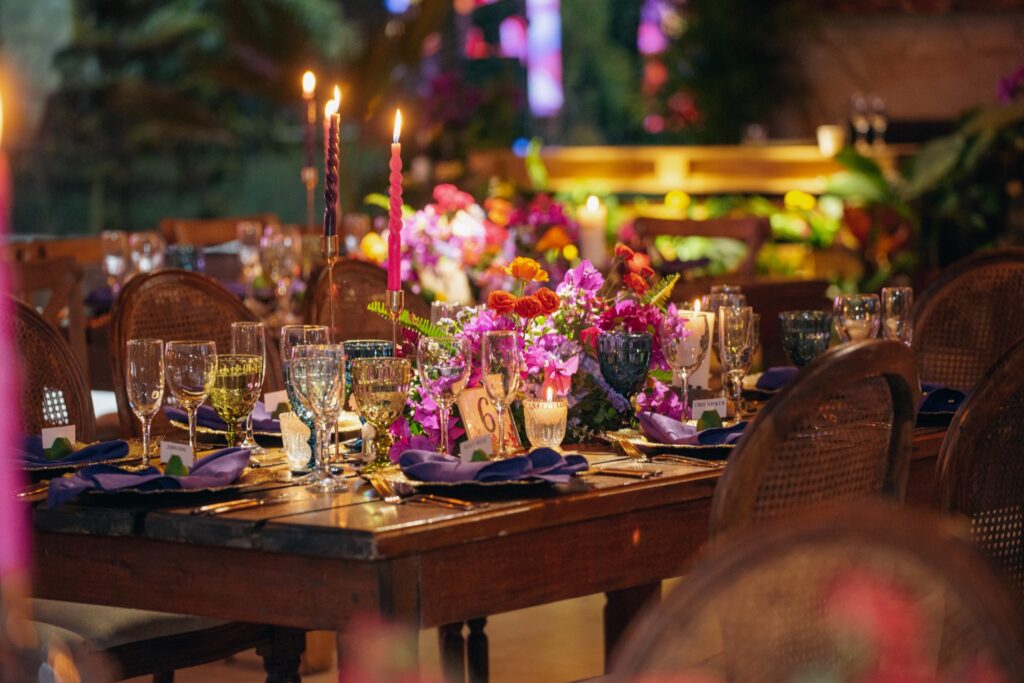 Centro de mesa de boda con flores coloridas en tonos morados, fucsias y naranjas, copas en vidrio de colores y candelabros con velas altas en una mesa de madera