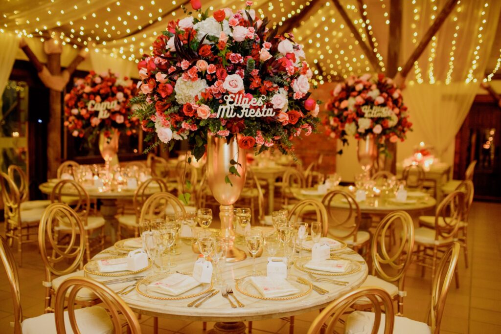 Centro de mesa de boda elevado en copa dorada con flores en tonos rojos, rosados y blancos, acompañado de sillas doradas y decoración con luces cálidas