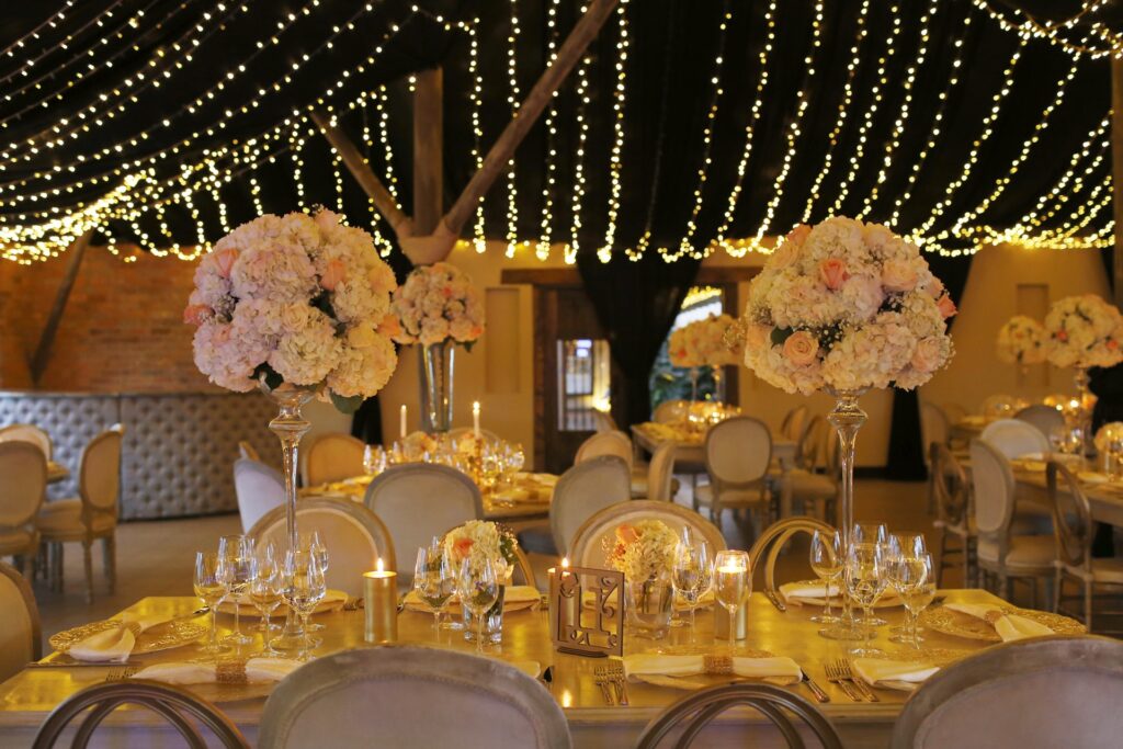 Centros de mesa altos con flores blancas y rosas en tonos nude, sobre copas de cristal, con decoración de boda iluminada por guirnaldas de luces