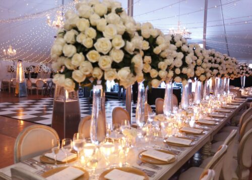 Centros de mesa con rosas blancas en copas altas de cristal, decoración elegante de boda con mesa larga iluminada por velas y guirnaldas de luces
