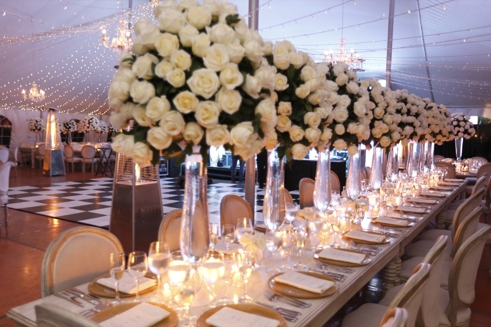 Centros de mesa con rosas blancas en copas altas de cristal, decoración elegante de boda con mesa larga iluminada por velas y guirnaldas de luces
