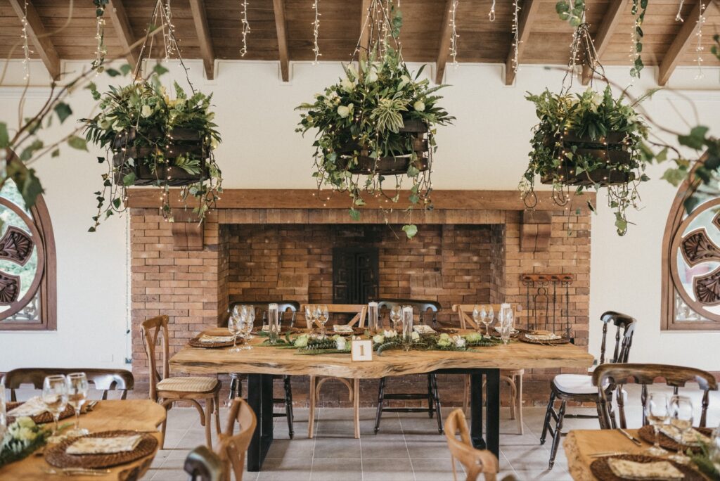 Decoración de boda rústica con centros de mesa de follajes verdes, velas en cilindros de cristal y arreglos colgantes naturales iluminados con luces cálidas