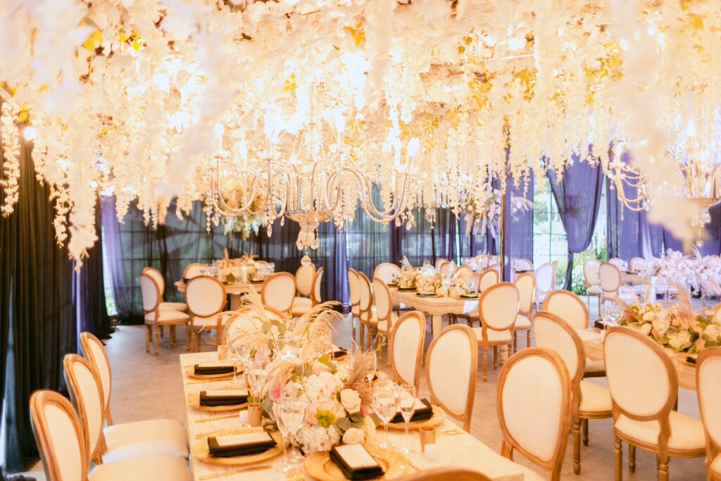 Decoración de boda con techo cubierto de flores blancas colgantes, candelabros de cristal y mesas elegantes con sillas estilo Luis XVI en tonos claros.